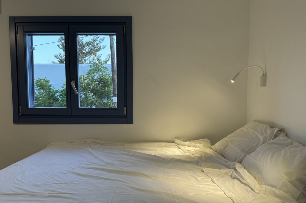 Υπνοδωμάτιο με λευκά σεντόνια και μπλε παράθυρο σε ανακαίνιση κατοικίας στην Τήνο από το SCale Design Studio.