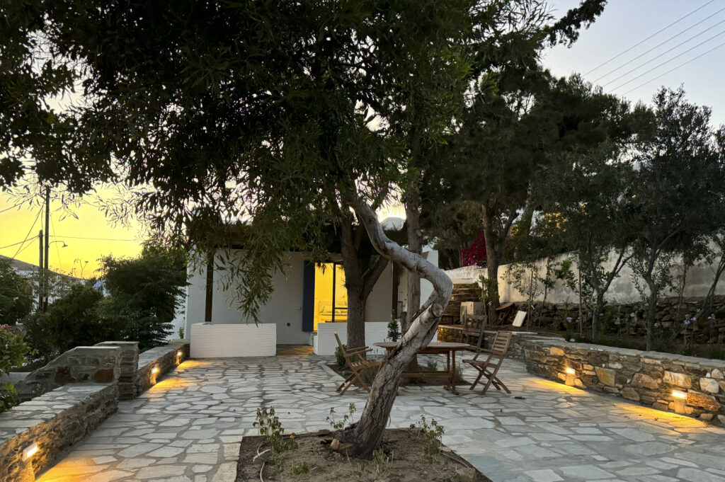 Πέτρινη αυλή κατοικίας στην Τήνο με εξωτερικό φωτισμό και τραπεζαρία κάτω από δέντρο από το SCale Design Studio.